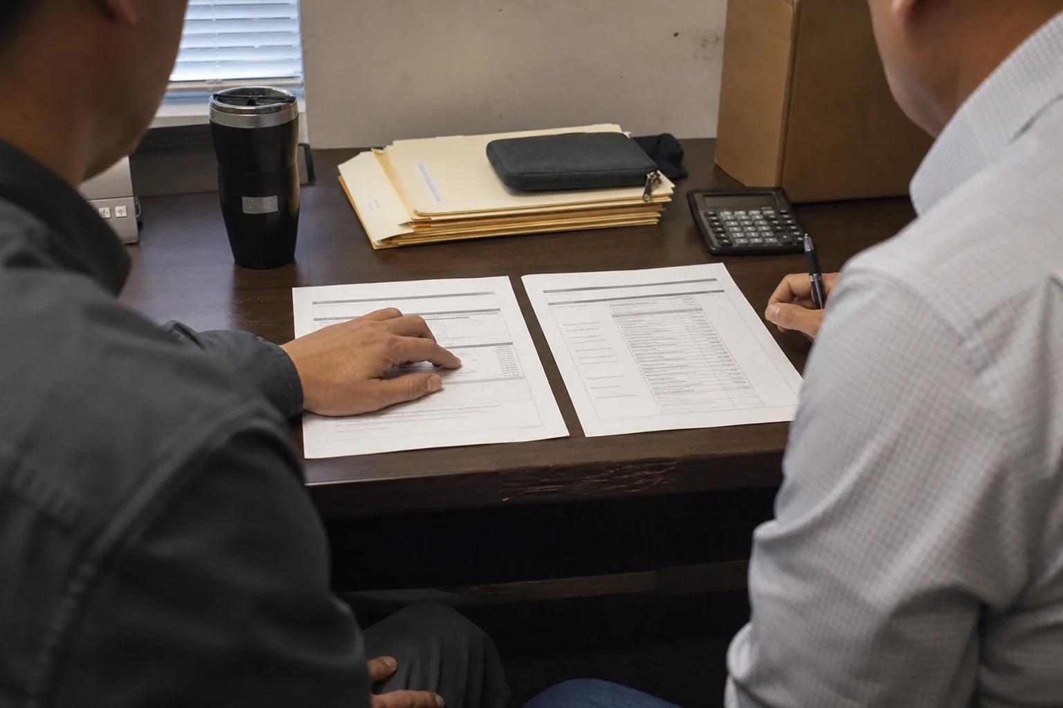 Over-the-shoulder view of two men at an office desk reviewing two printed documents side by side, with manila folders, a calculator and a travel mug nearby