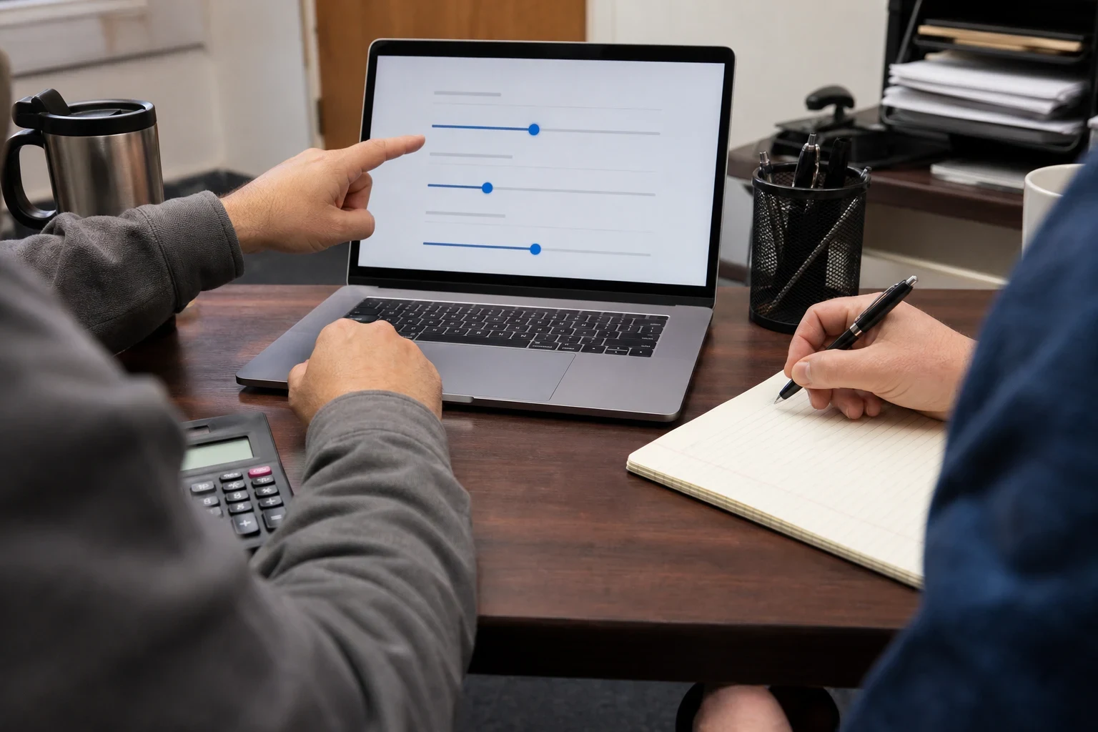Two people at a wooden office desk; one points at adjustable sliders on a laptop screen while the other writes notes on a lined notepad beside a calculator.